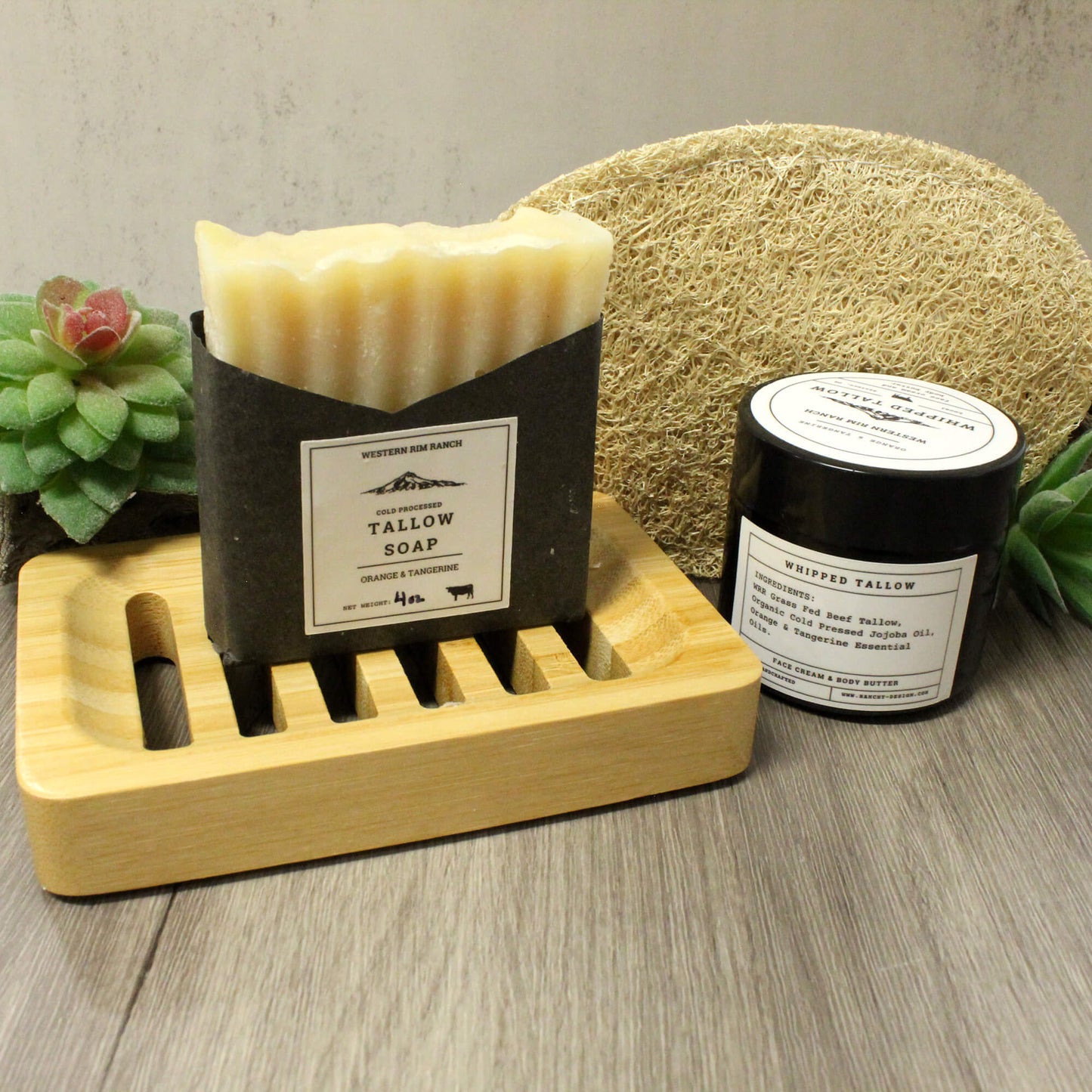 A bar of handmade soap in a black paper sleeve and a small black jar with a white label, styled on a wooden soap dish with a natural loofah and succulents in the background.