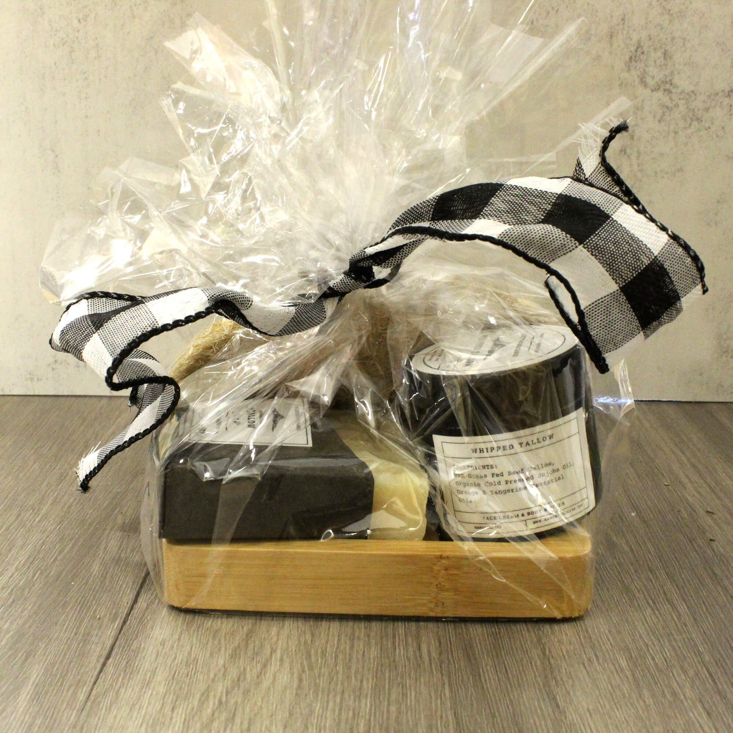 A gift set in clear cellophane wrapping with a black-and-white gingham ribbon, containing a dark soap bar, a cream-colored bar, and a small jar with a white label, displayed on a wooden tray over a wood surface.