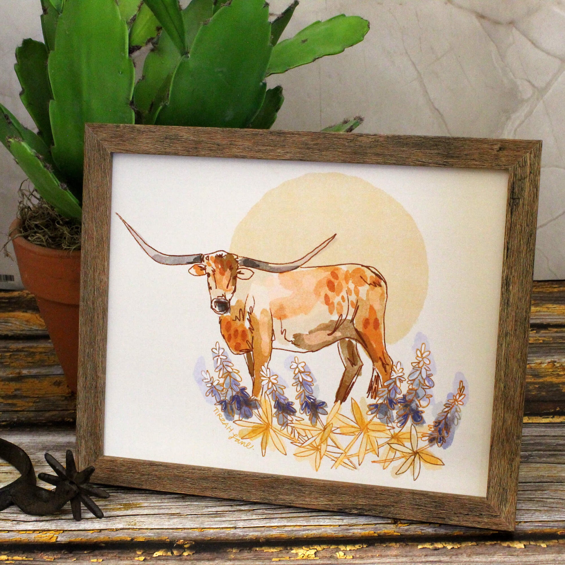 Framed artwork of a longhorn cow with flowers on a wooden surface.