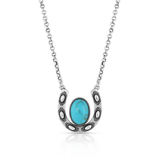 We feel lucky to wear this pendant necklace, inspired by horseshoes and the blue of the Big Sky above us. It is silver-toned and smooth. The horseshow pendant is held from both tops to lay flat. it has punched rivets and detailed etching for character. Within the center is an oval turquoise stone. It hangs from a 16" chain.