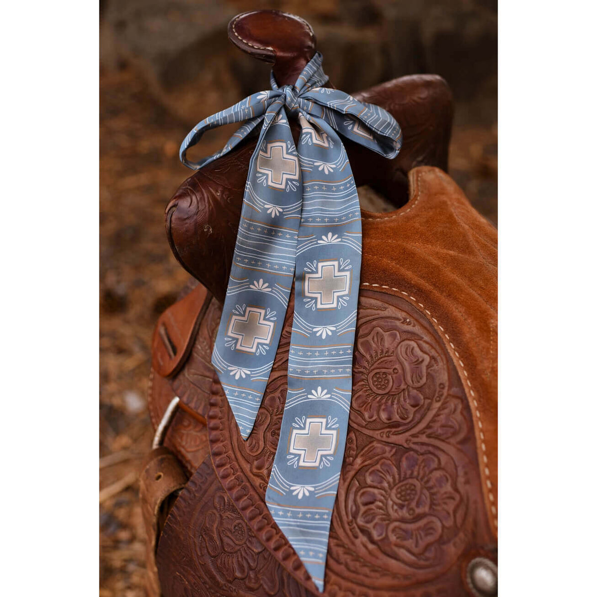A light blue patterned silk scarf with white crosses and western-style stripes, tied around the saddle of a brown tooled leather western saddle.