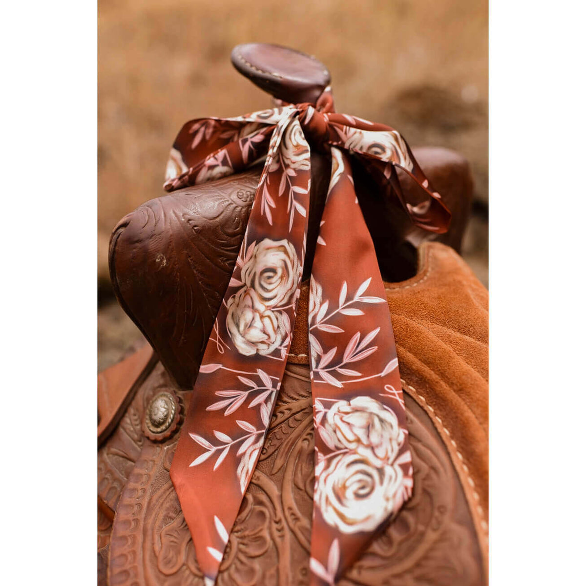 A rust-orange satin scarf with a white rose and leaf pattern tied around the saddle of a brown leather western saddle.