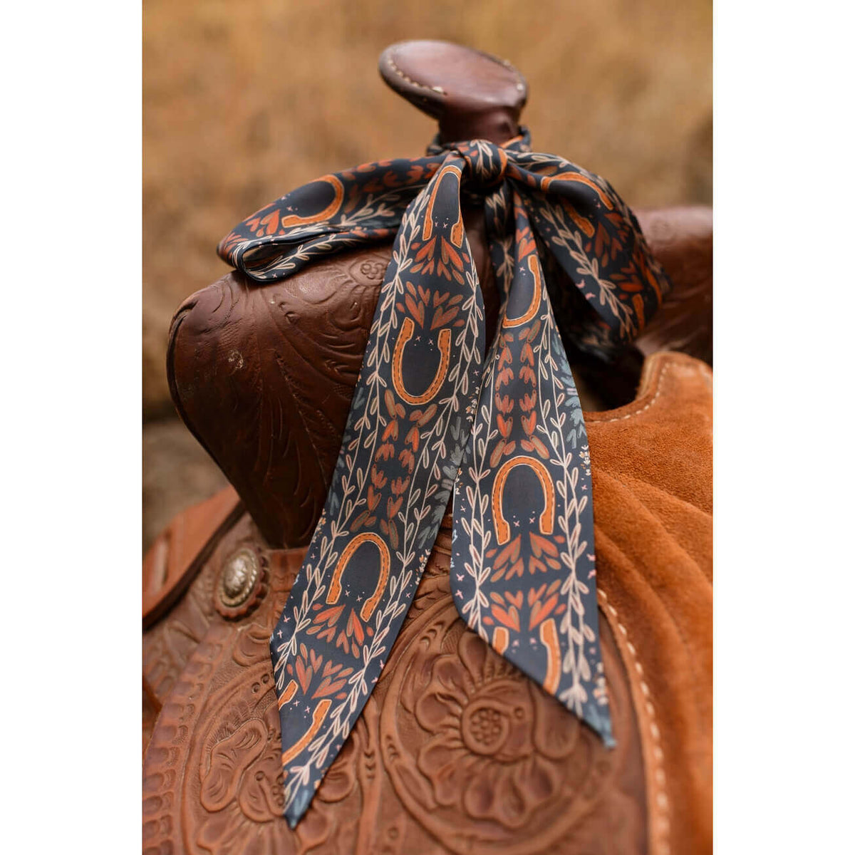 A patterned silk twilly tied around a brown leather western saddle, featuring a design of orange horseshoes and leafy motifs on a dark gray background.