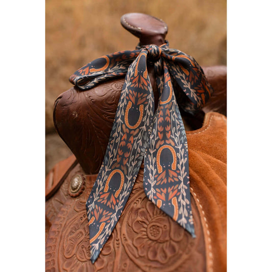 A patterned silk twilly tied around a brown leather western saddle, featuring a design of orange horseshoes and leafy motifs on a dark gray background.