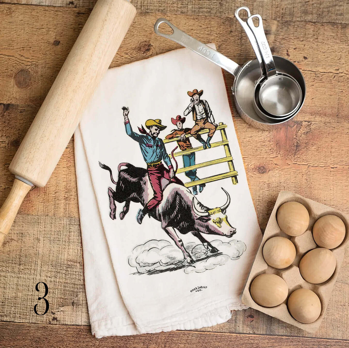 A kitchen towel with a colorful illustration of a cowboy riding a bull, styled on a rustic wooden table with a wooden rolling pin, metal measuring cups, a carton of eggs, and a wooden spoon.