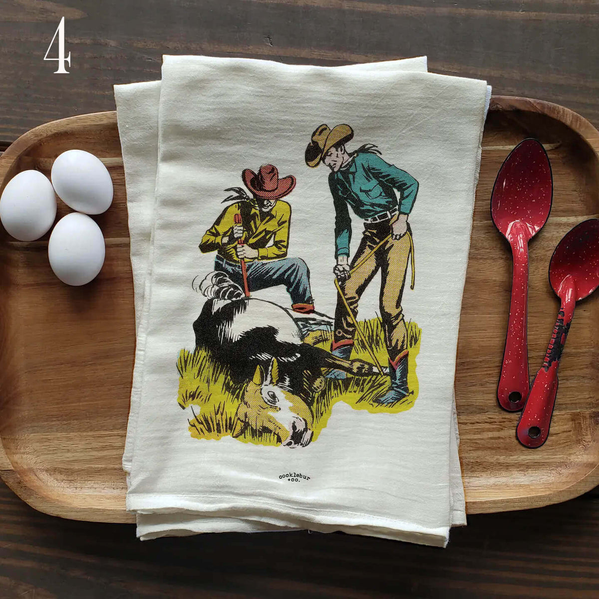 A stack of cream-colored kitchen tea towels with a vintage cowboy illustration, displayed on a wooden serving tray alongside three white eggs and two red speckled spoons.
