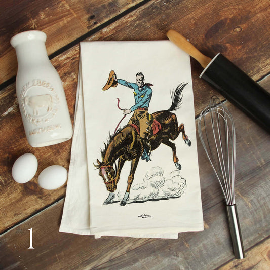 A white kitchen tea towel with a western cowboy riding a bucking horse illustration, styled on a rustic wooden table with baking props including a vintage-style milk bottle, eggs, a rolling pin, and a whisk.