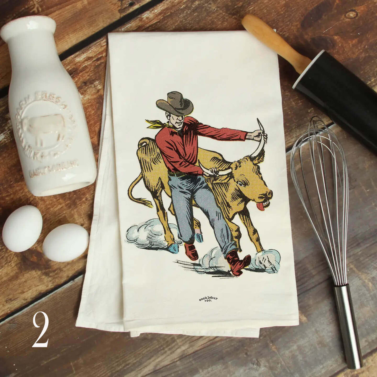 A stack of white kitchen towels featuring a vintage-style illustration of a cowboy riding a calf, styled on a rustic wooden table with a white milk bottle, eggs, rolling pin, and whisk.