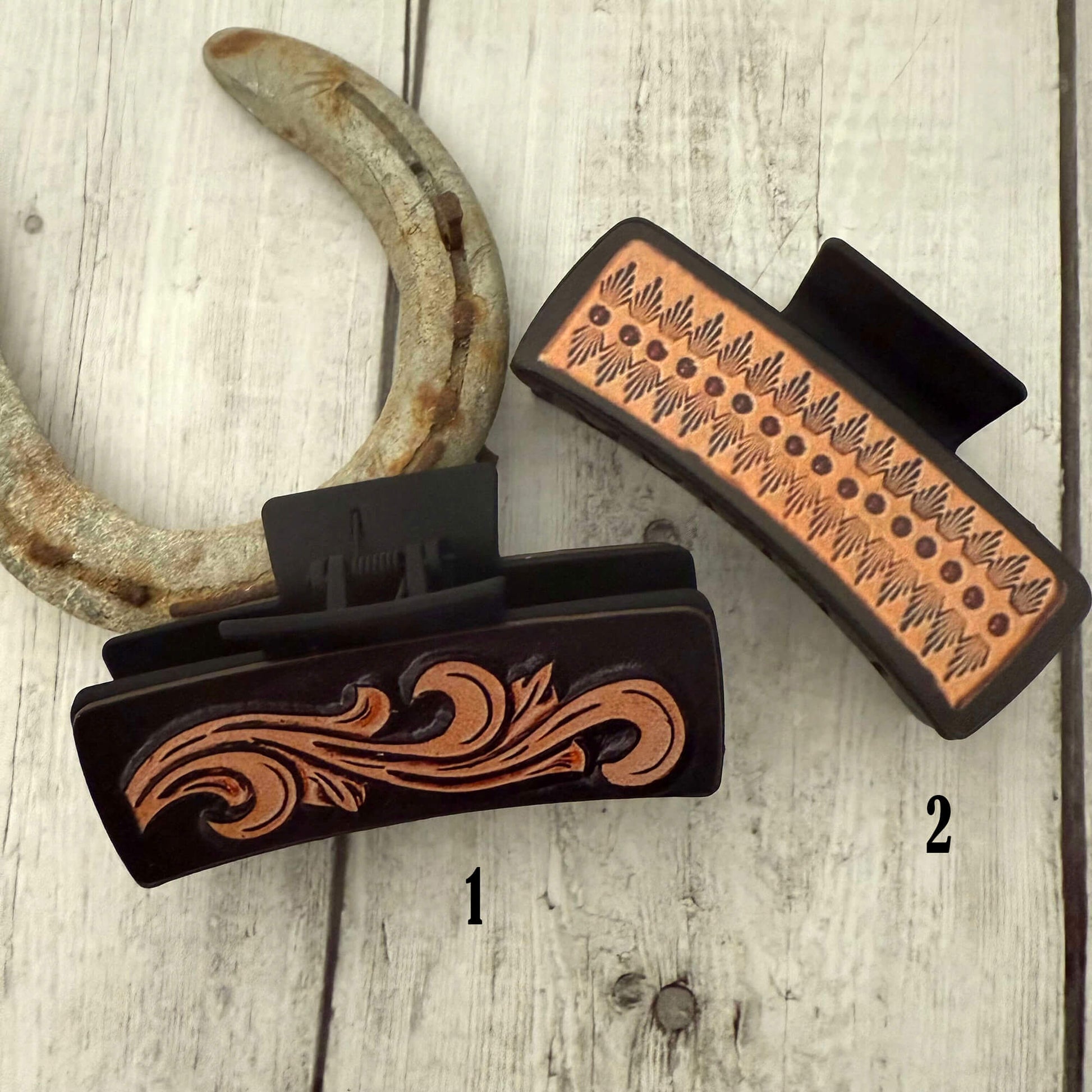Two hair clips with embossed designs on a wooden surface, one of which is attached to a rusty horseshoe.