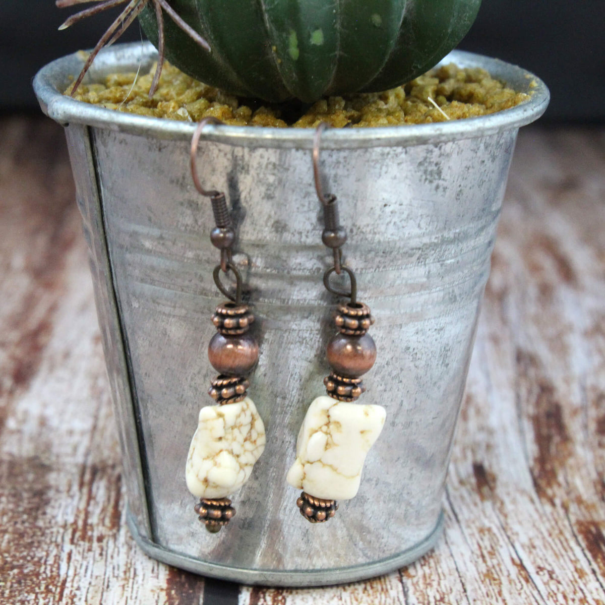 A pair of dangling earrings with cream-colored cowrie shells and bronze-toned beads, turquoise bead hanging from a galvanized metal planter pot on a rustic wooden surface.