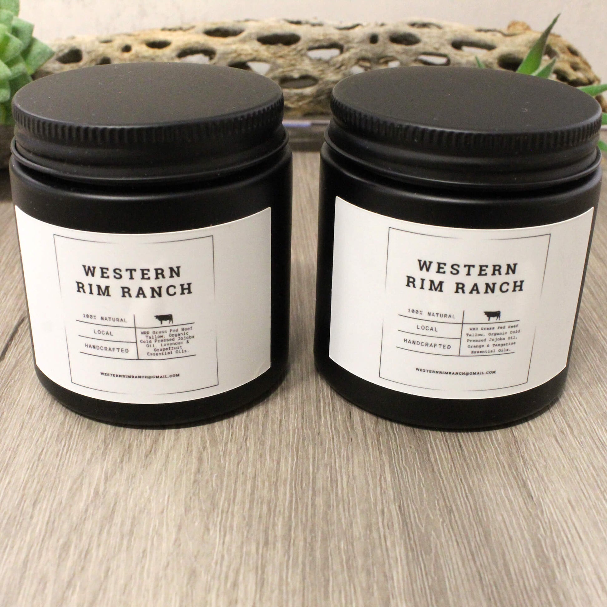 Two black plastic jar candles with white labels reading 'WESTERN RIM RANCH' sit on a light wood surface in front of a woven decorative mat.