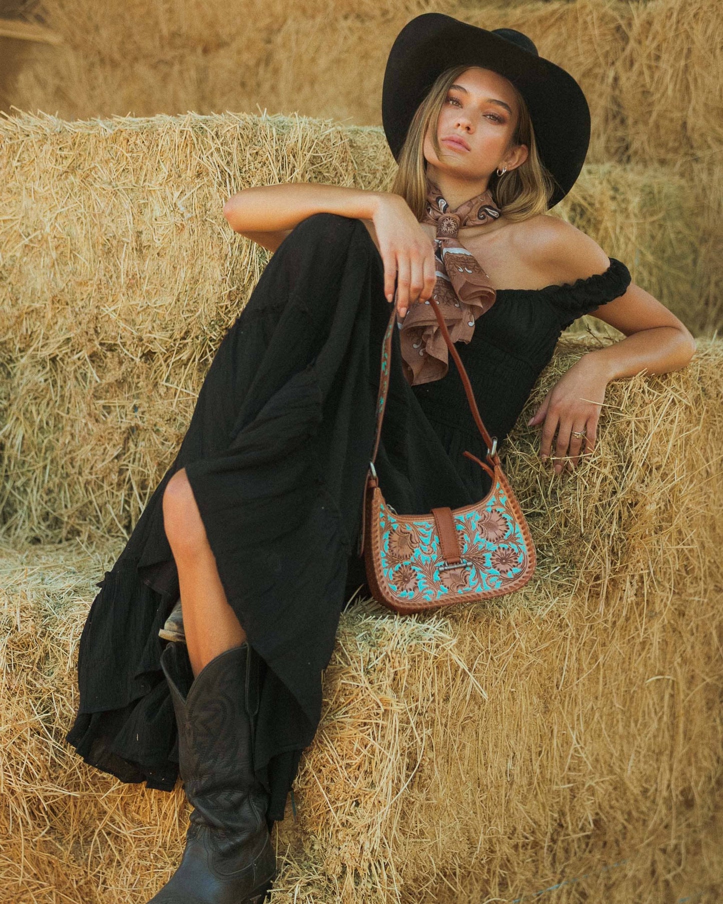 Woman in black dress and hat sitting on hay with a patterned handbag