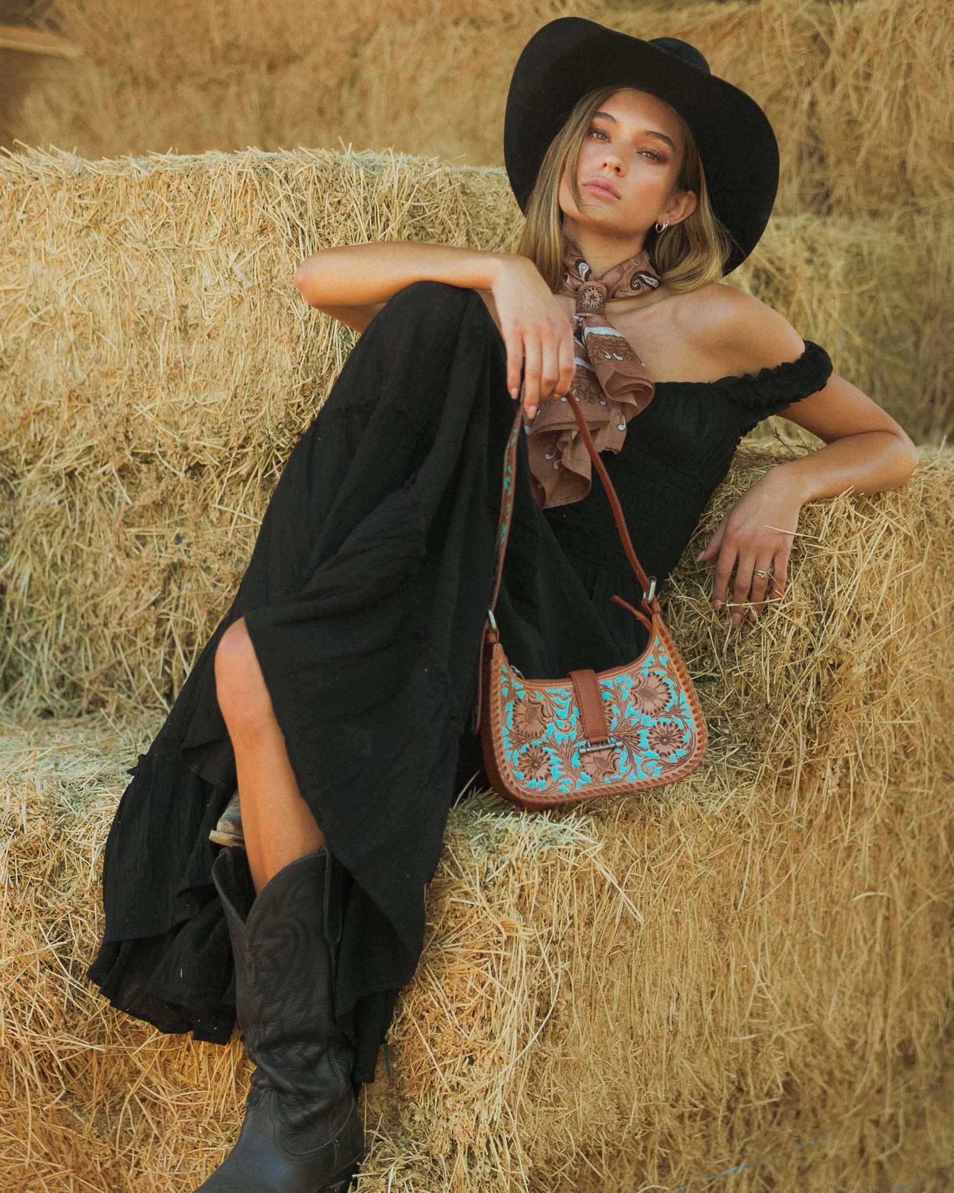 Woman in black dress and hat sitting on hay with a patterned handbag