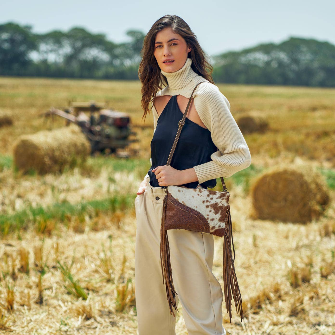 Woman standing in a field with hay bales, holding a cowhide bag.