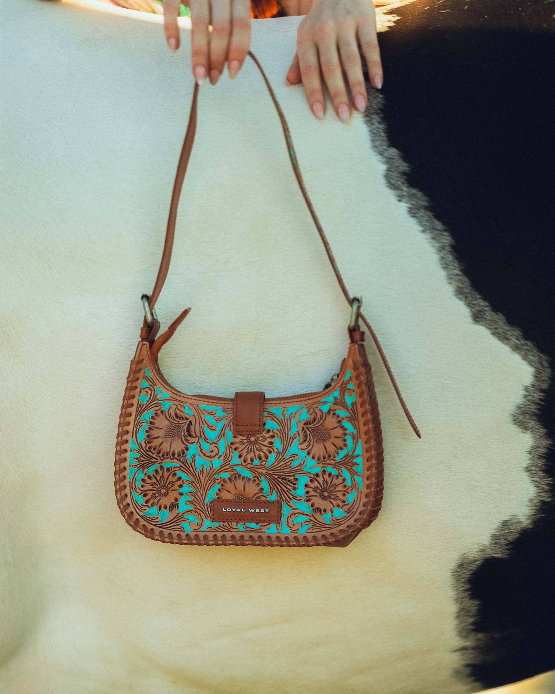 Brown leather handbag with teal embroidery held by a person on a white background