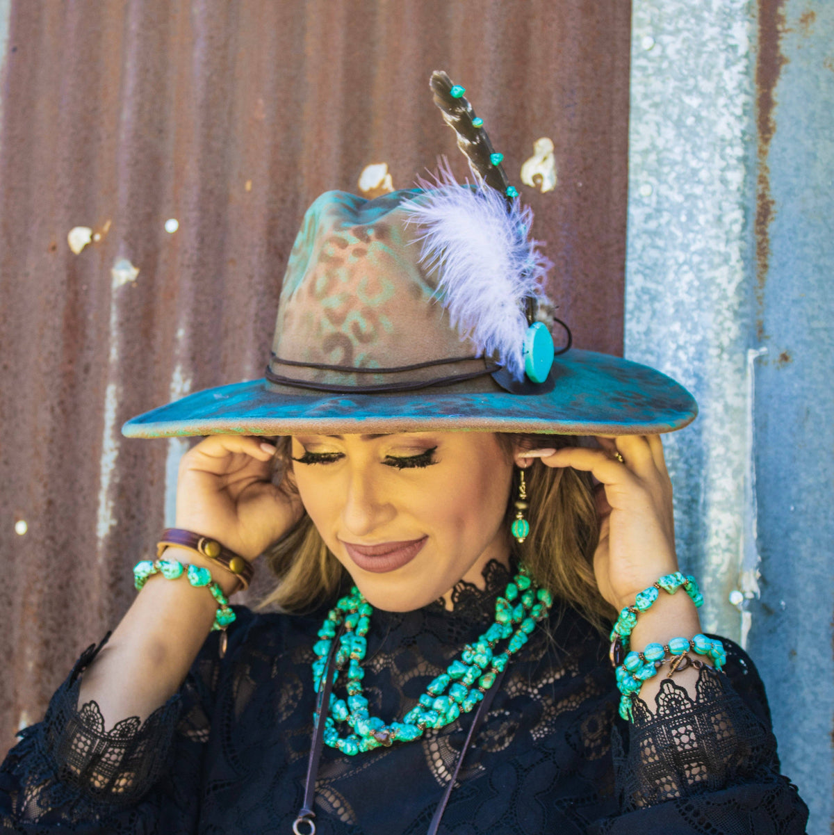 Woman wearing a decorative hat with feathers and turquoise jewelry against a rustic wall.