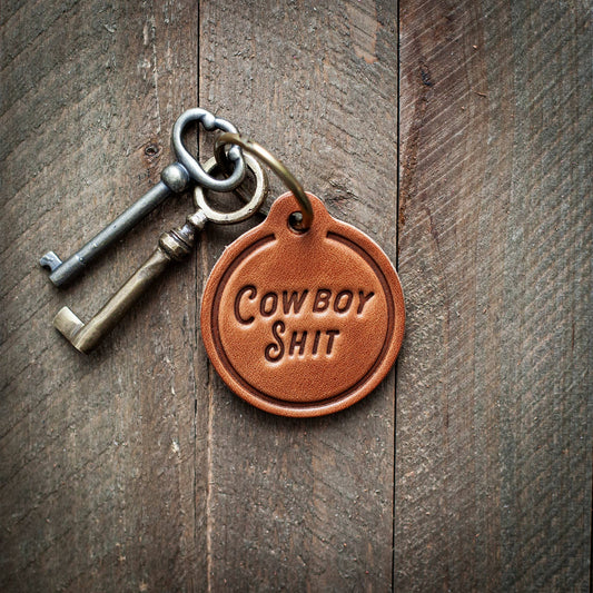 Keychain with 'Cowboy Shit' tag on a wooden surface