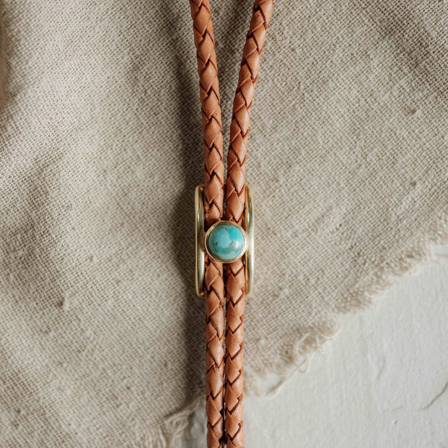 Brown braided leather bolo tie with a turquoise stone on a textured beige background
