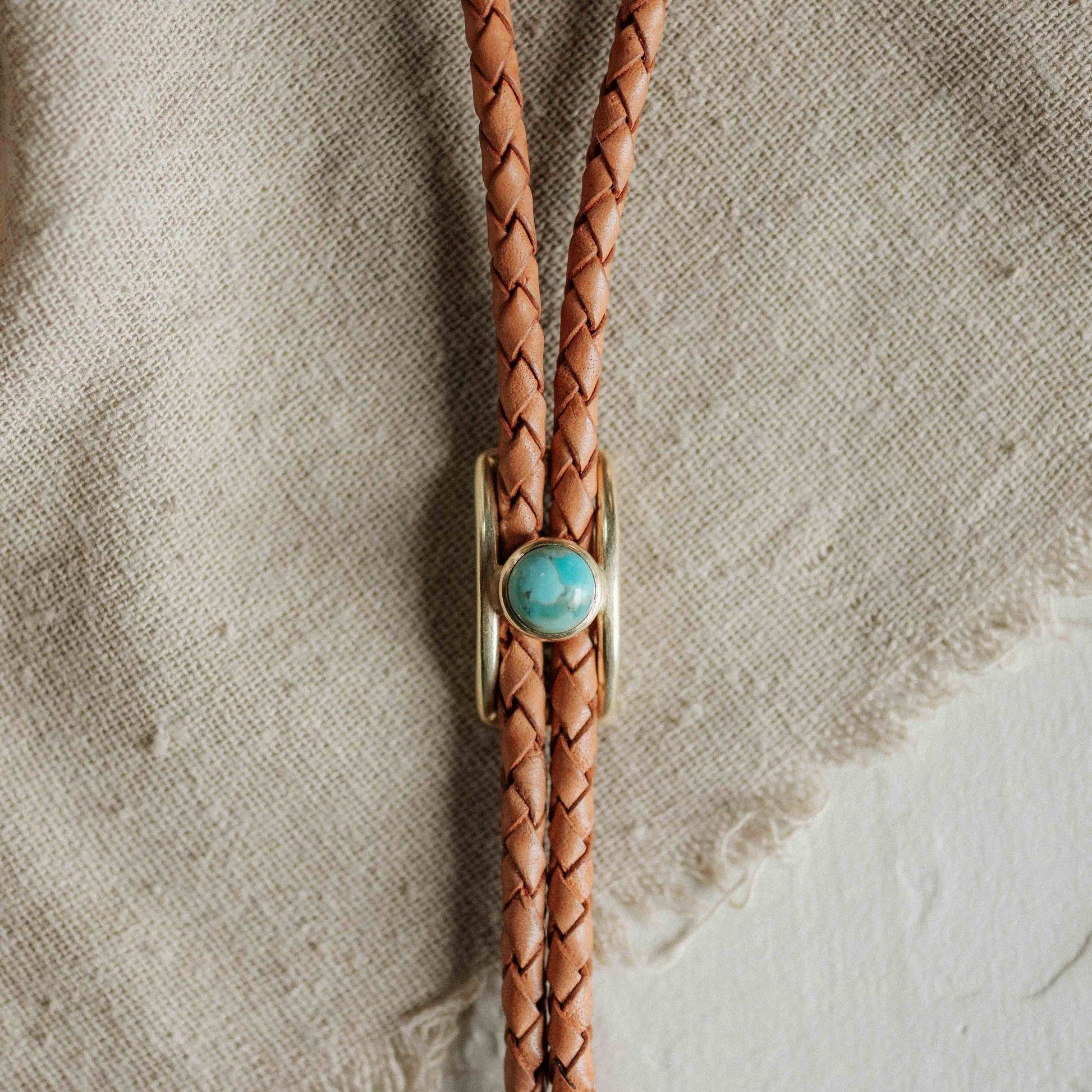 Brown braided leather bolo tie with a turquoise stone on a textured beige background